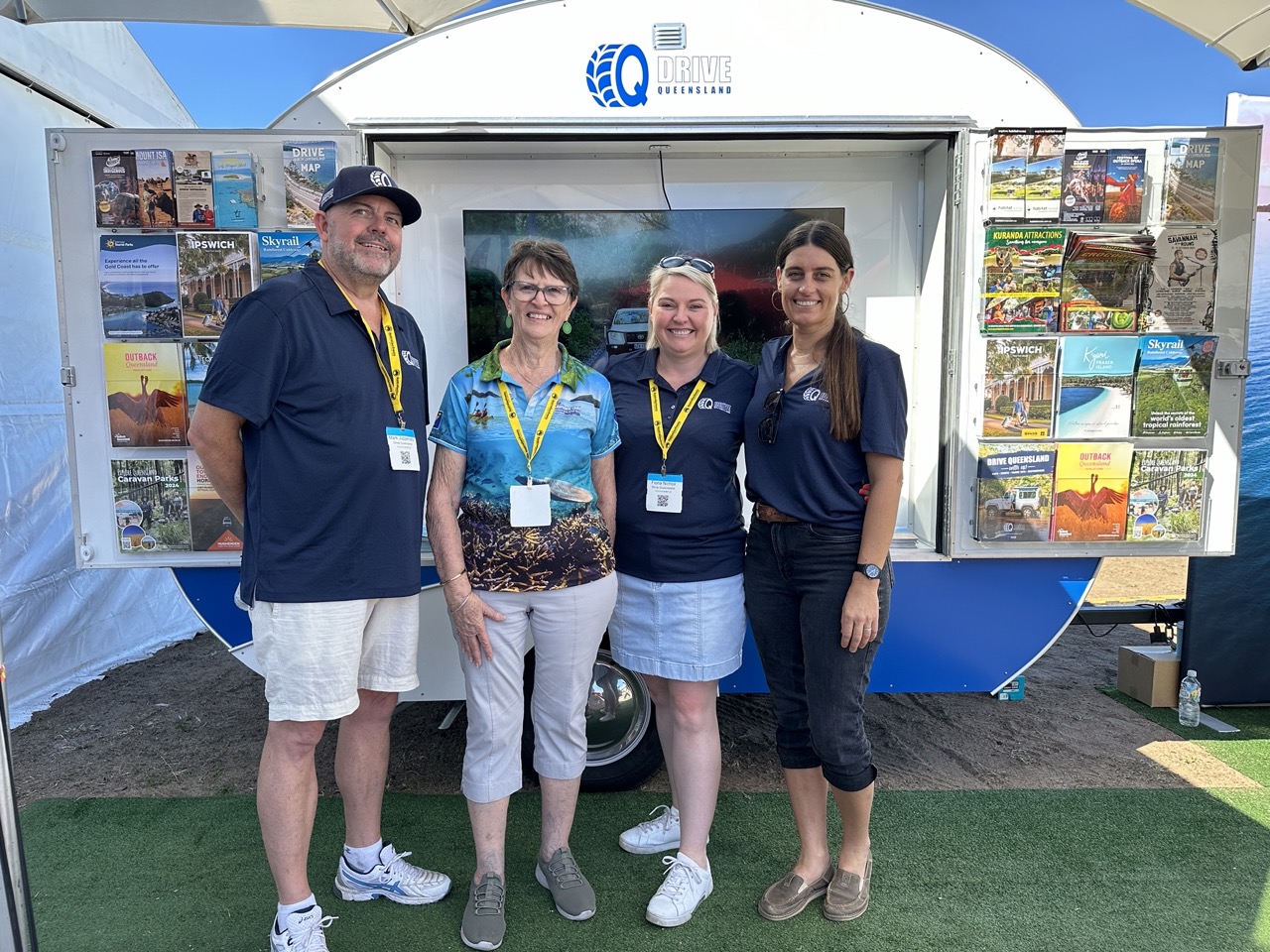 Four people stand in front of a display trailer with brochures. They are wearing casual shirts and name tags, smiling for the camera. The trailer has a logo on it.