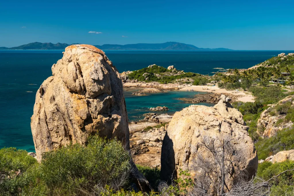 Scenic view of a coastal landscape with large rock formations in the foreground, turquoise water, and distant mountains under a clear blue sky.
