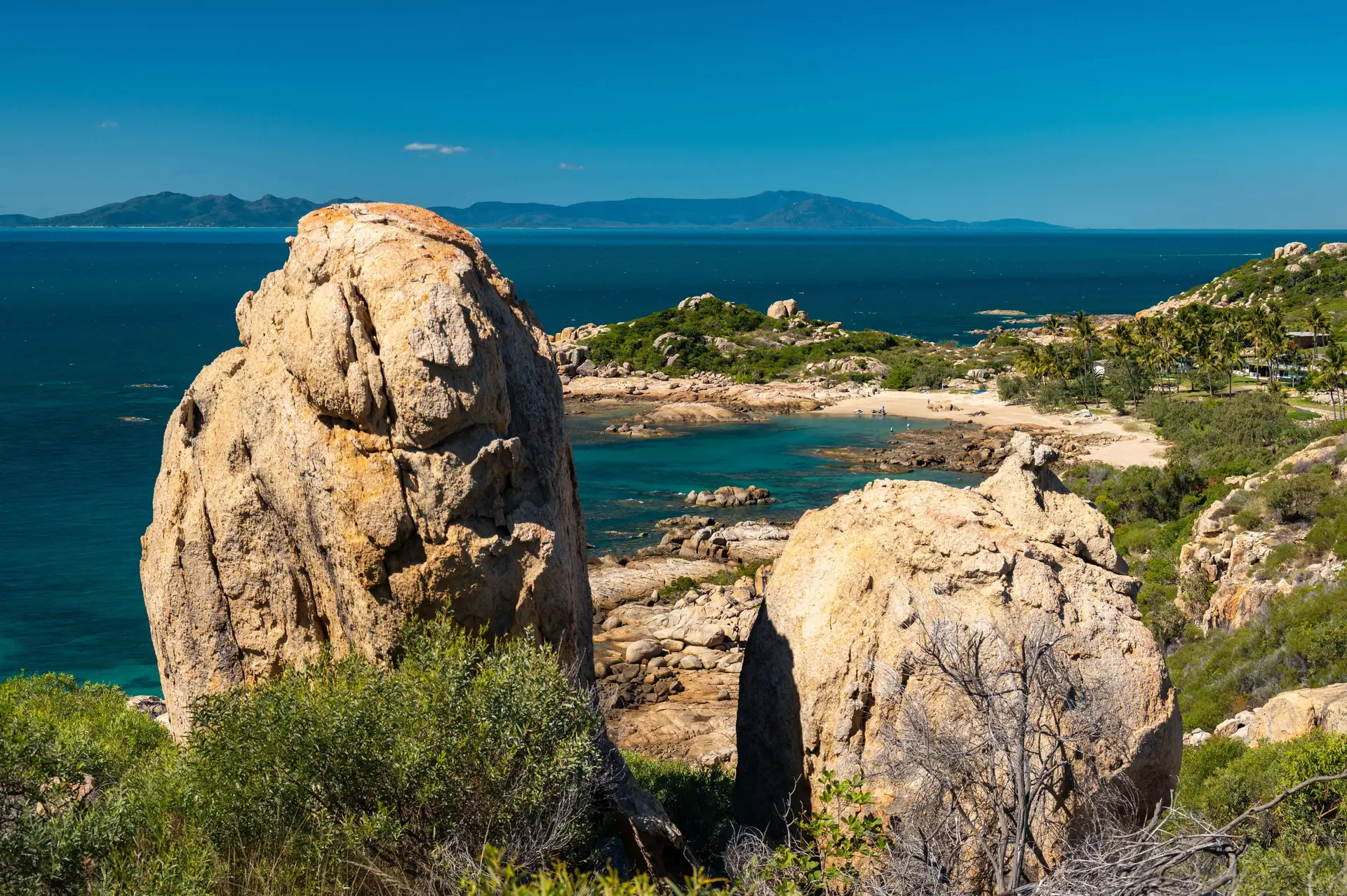 Scenic view of a coastal landscape with large rock formations in the foreground, turquoise water, and distant mountains under a clear blue sky.