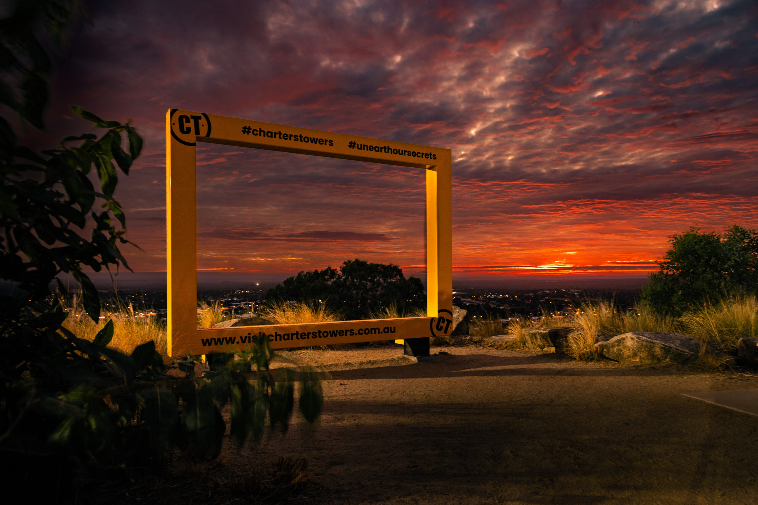Large yellow picture frame structure overlooks a vibrant sunset sky with scattered clouds and city lights in the distance, surrounded by grass and rocks.