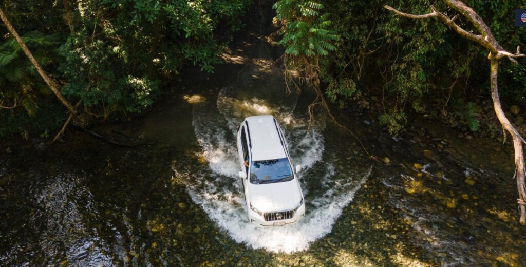 A white SUV drives through a shallow, rocky stream surrounded by dense green forest.