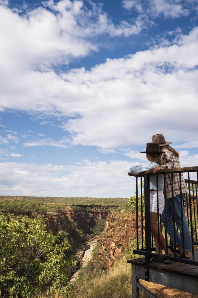 Two people in casual clothing and hats stand at an overlook railing, gazing at a scenic canyon under a partly cloudy sky.