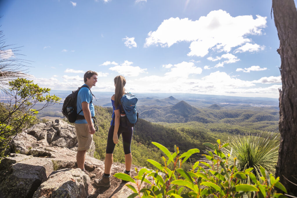 Two hikers with backpacks stand on a rocky mountain overlook, gazing at a vast, forested landscape under a partly cloudy sky.