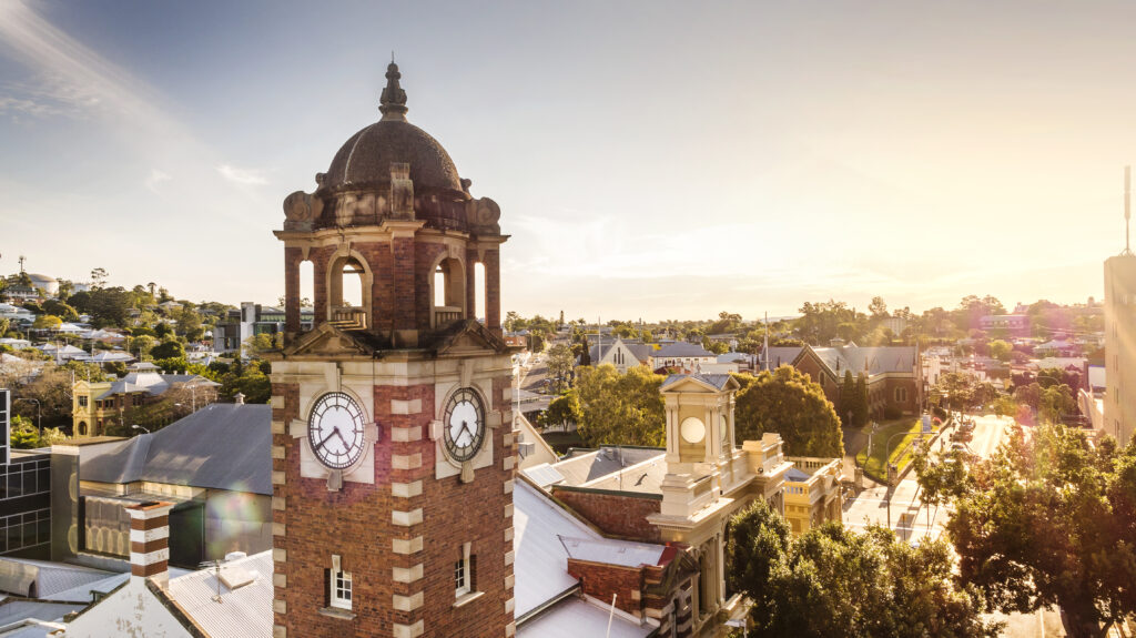 A brick clock tower stands tall against a backdrop of a cityscape with trees, buildings, and a bright sunset.