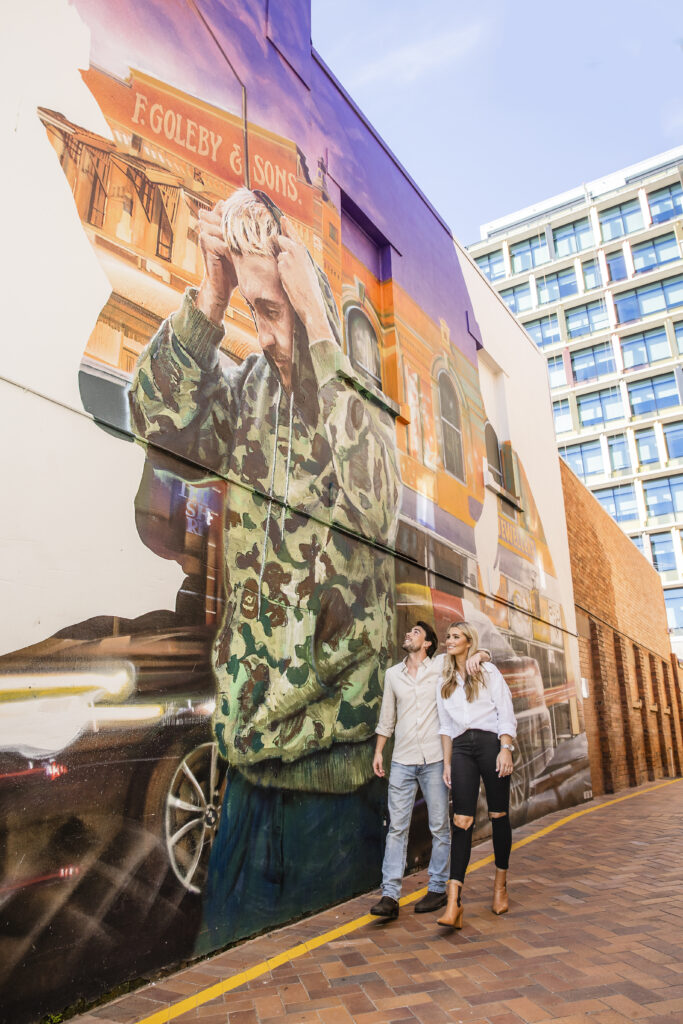 Two people walk past a large mural depicting a person in a camo jacket adjusting their hair. The mural is on a brick wall next to a modern building.
