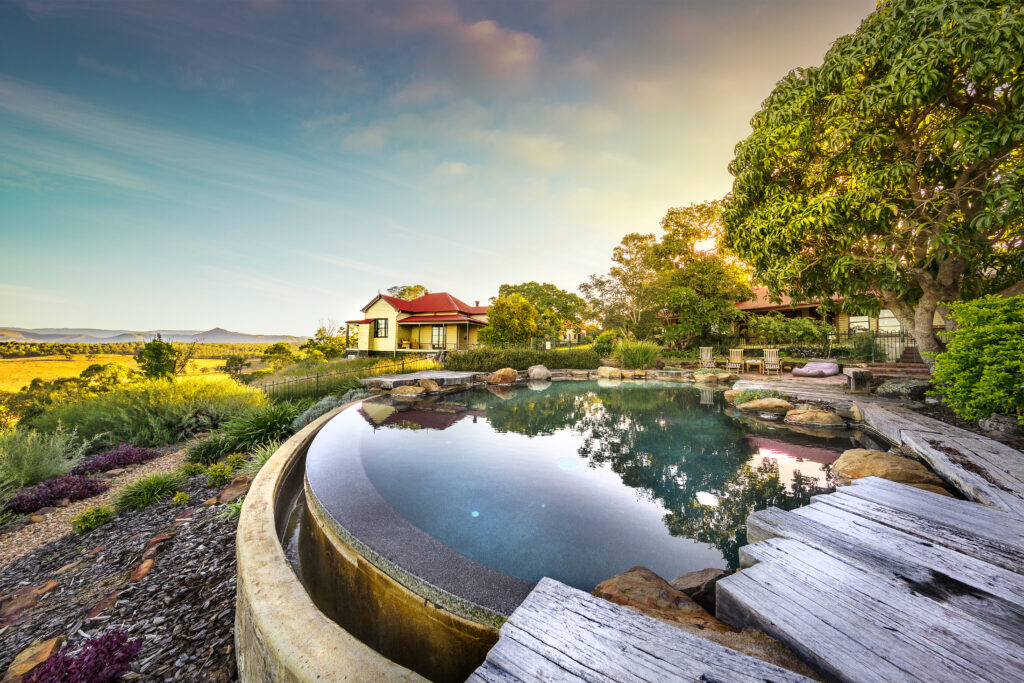 A scenic view of a round infinity pool overlooking a lush landscape with few houses, trees, and hills in the background under a serene sky.