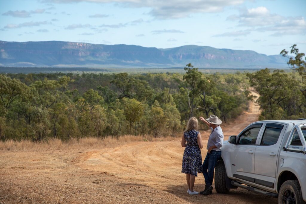 A man and woman stand by a white truck, looking at a scenic landscape with trees and distant mountains under a partly cloudy sky.