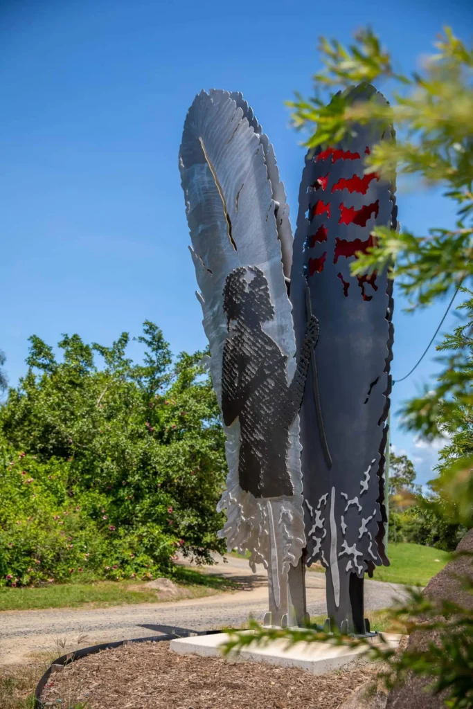 A metal sculpture depicts a silhouette of a person holding a feather with another feather beside it. The artwork stands outdoors with trees and greenery in the background.