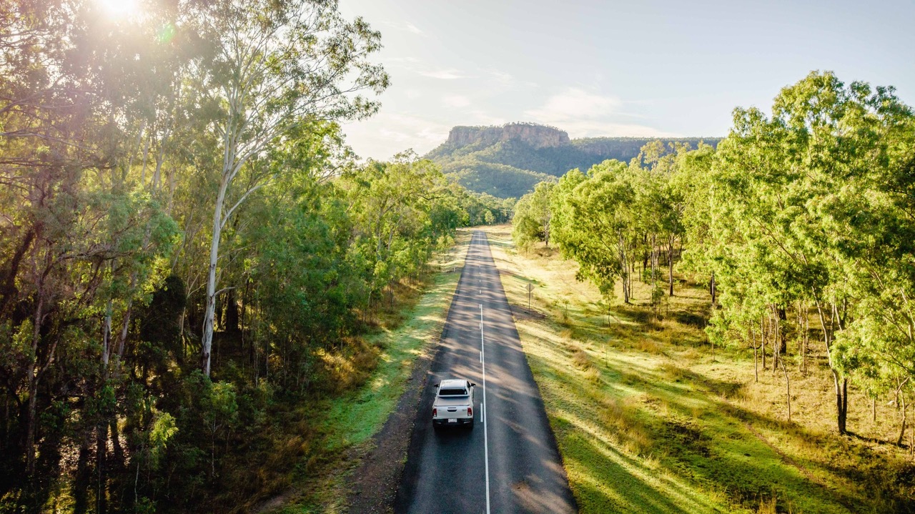A white car drives down a straight, tree-lined road surrounded by green hills and forests under a sunny sky.