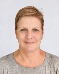 A person with short, light brown hair and a neutral expression, wearing a gray shirt, against a plain light background.