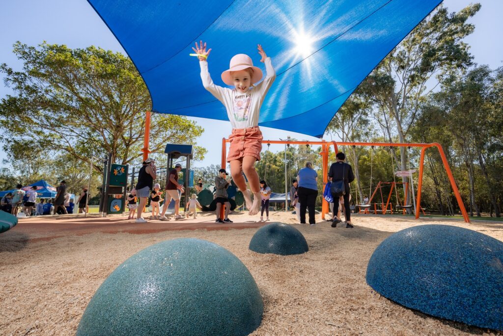 A child in a pink hat jumps off a playground feature under a blue sunshade, surrounded by people and trees.