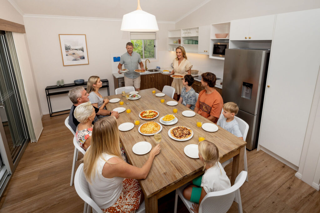 A family gathers around a wooden table in a modern kitchen, with adults serving food. Plates and orange-colored drinks are set for everyone.
