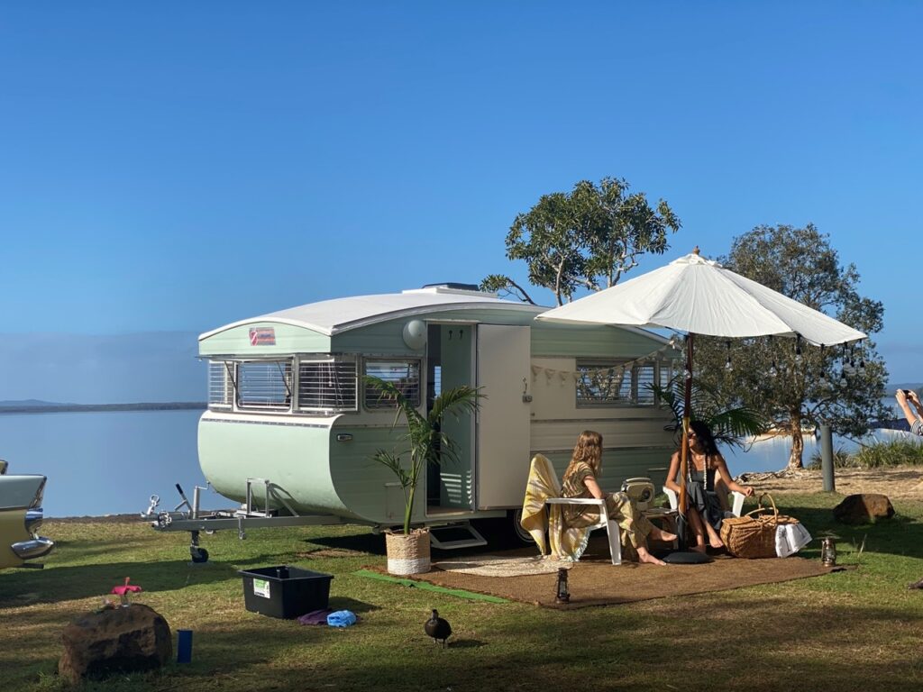 A vintage caravan is parked by a lake. Two people sit outside under an umbrella, surrounded by trees and grass. A duck stands nearby.