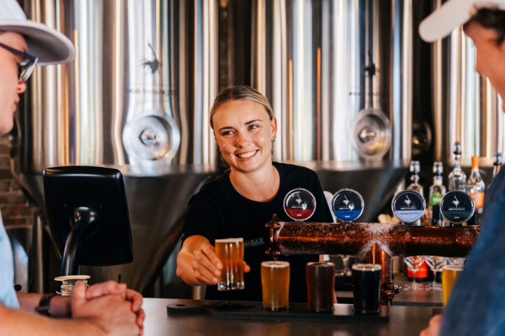 Bartender smiling while serving beer to a customer at a bar with large metal tanks in the background.