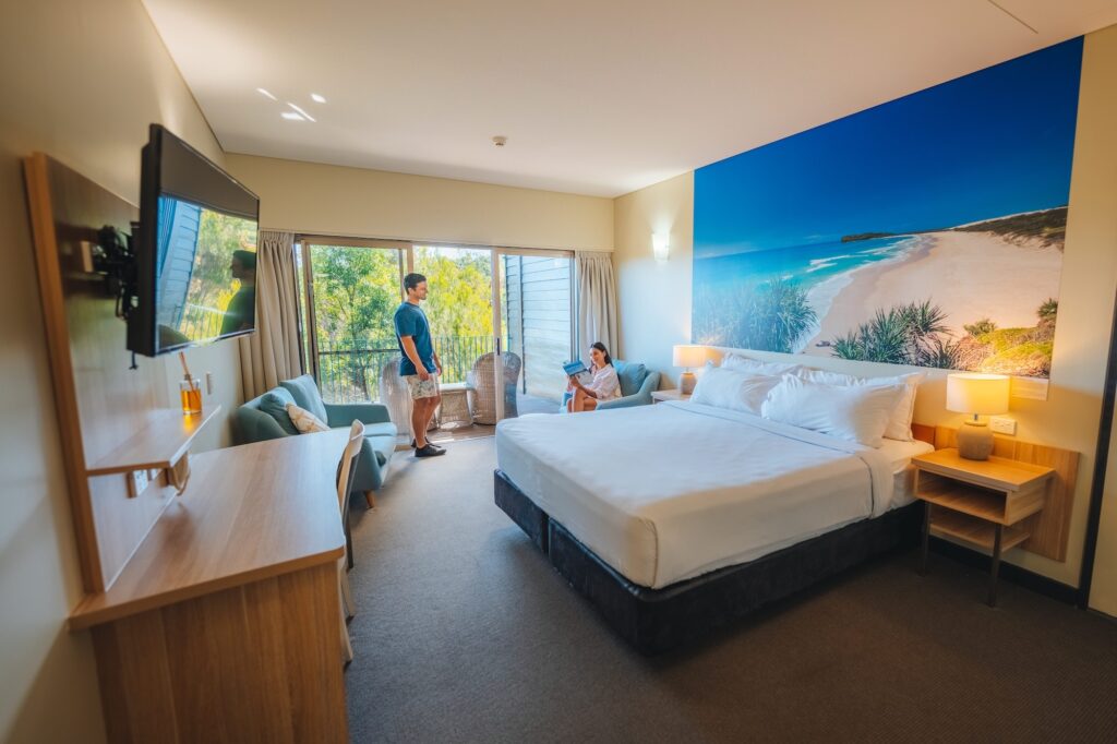 A hotel room with a large bed, a wall mural of a beach, a TV, seating area, and two people near the window overlooking greenery.