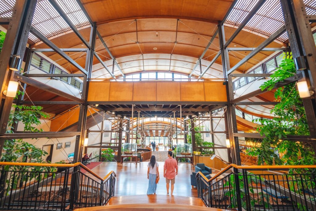 Two people walk inside a spacious, modern building with high wooden ceilings, large windows, metal beams, and abundant indoor plants.