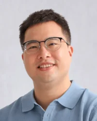 A man with short hair and glasses is smiling and wearing a blue collared shirt against a plain background.