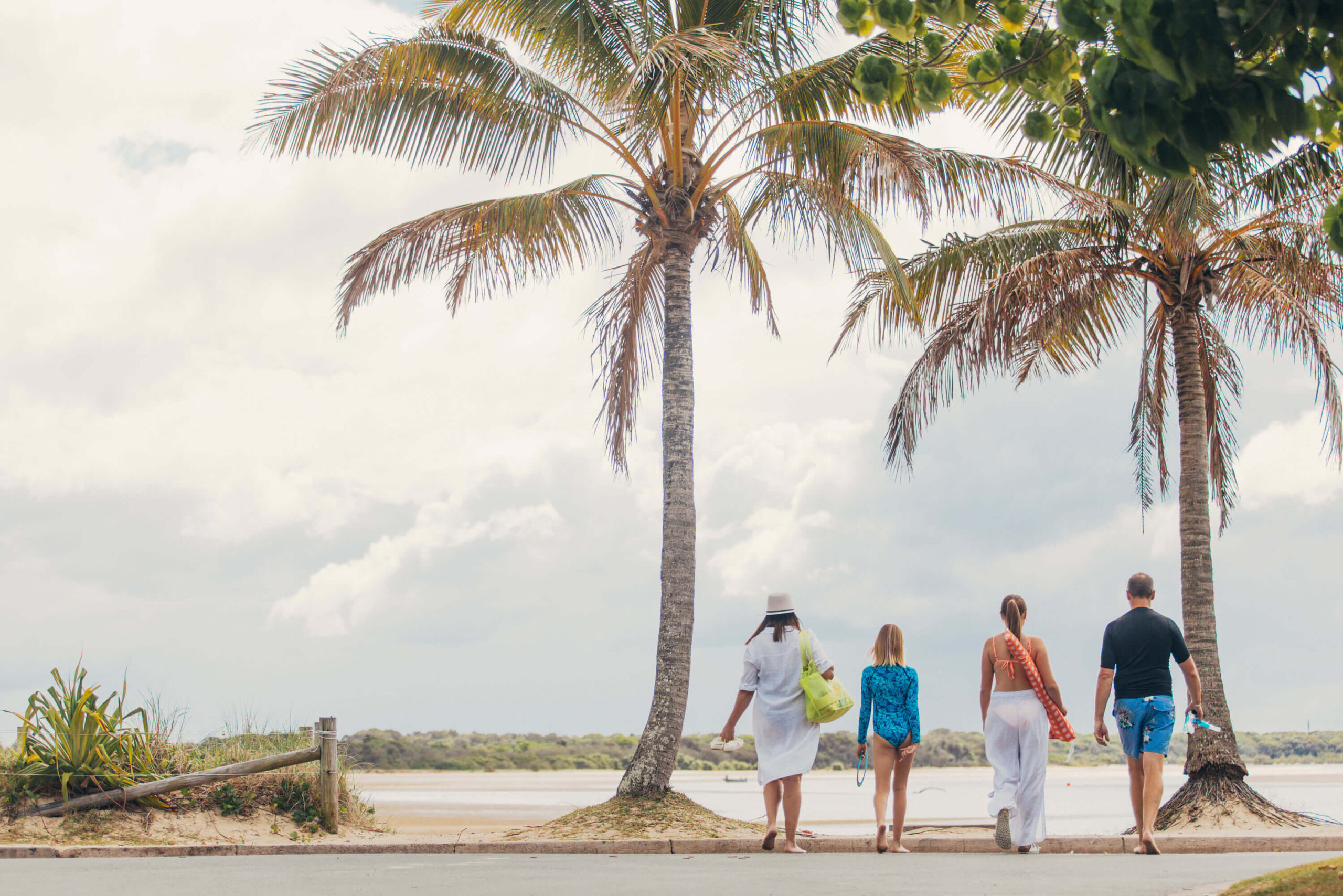 Four people walk toward the beach between two palm trees on a cloudy day.