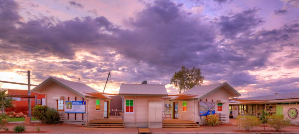 Small, colourful buildings under a cloudy sunset sky, with a large sign on the left and a tree in the background.