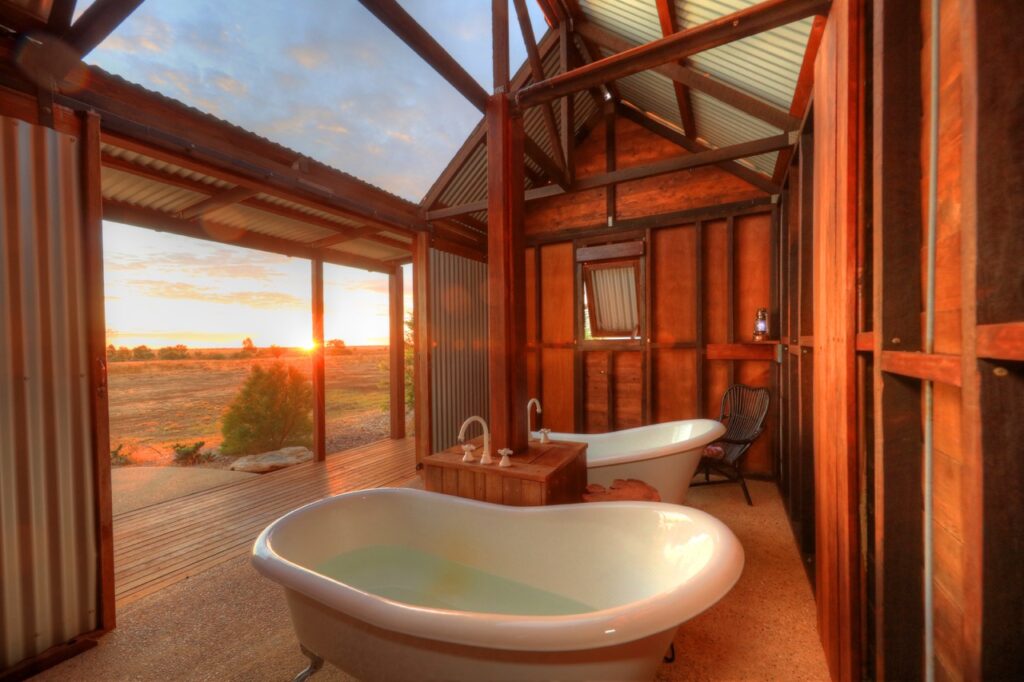 Two freestanding bathtubs in a rustic outdoor bathroom with wooden walls and a metal roof, overlooking a sunset view.