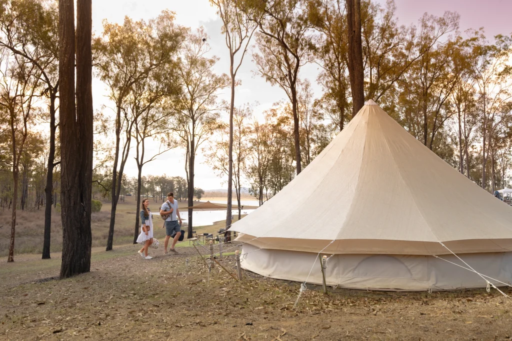 A couple walking near a large beige tent in a wooded area during sunset.