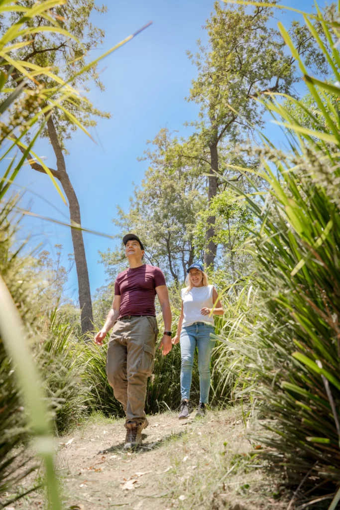 Two people walking on a grassy trail surrounded by tall trees and plants, under a clear blue sky.