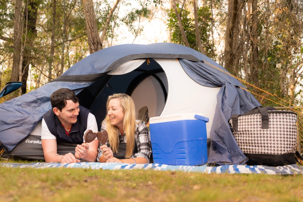 A man and woman lie on a blanket in front of a tent, holding ice cream bars. A cooler and a basket are nearby. The scene is set in a wooded area.