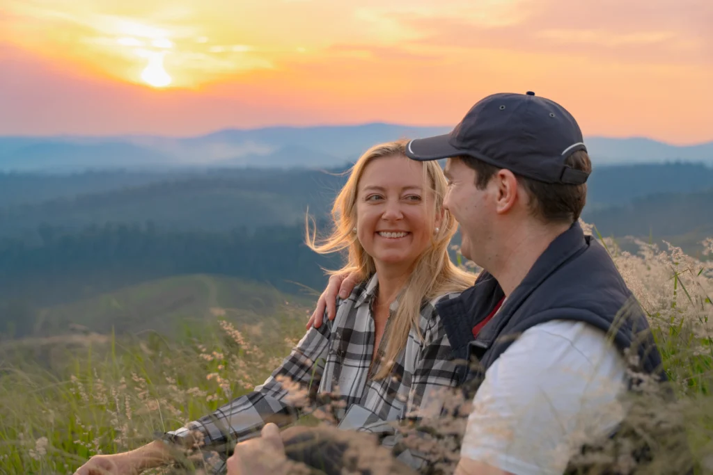A couple sits on grass, smiling at each other as the sun sets over a mountainous landscape.
