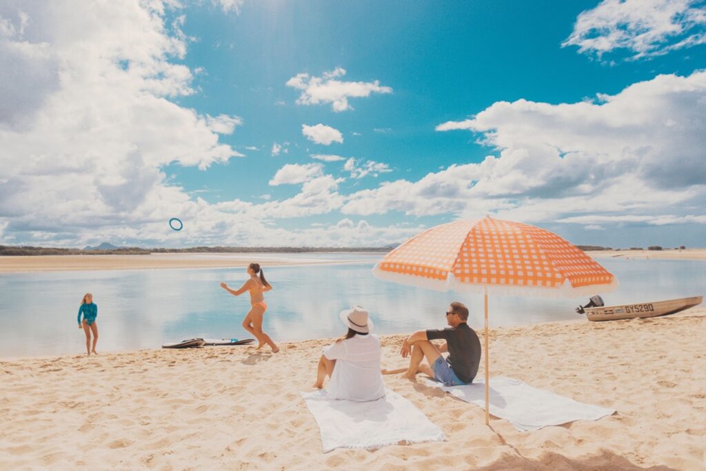 People relaxing on a sandy beach under a blue sky. Two people are sitting under an orange umbrella, while two others are playing near the water. A boat is visible in the background.
