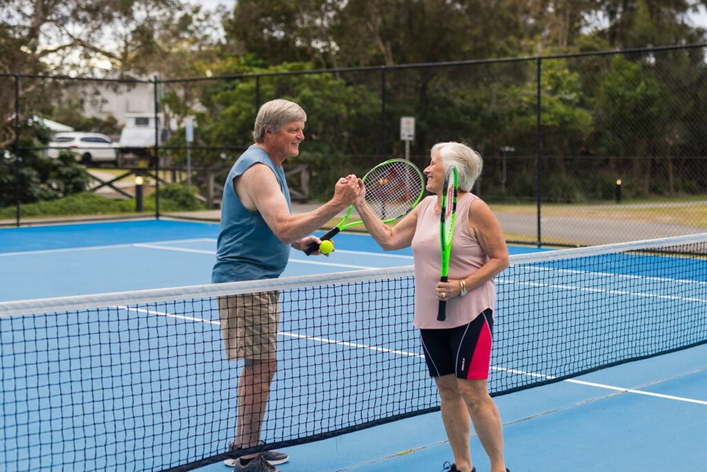 Two people on a tennis court exchange a fist bump over the net while holding tennis rackets. They are on an outdoor court with trees in the background.