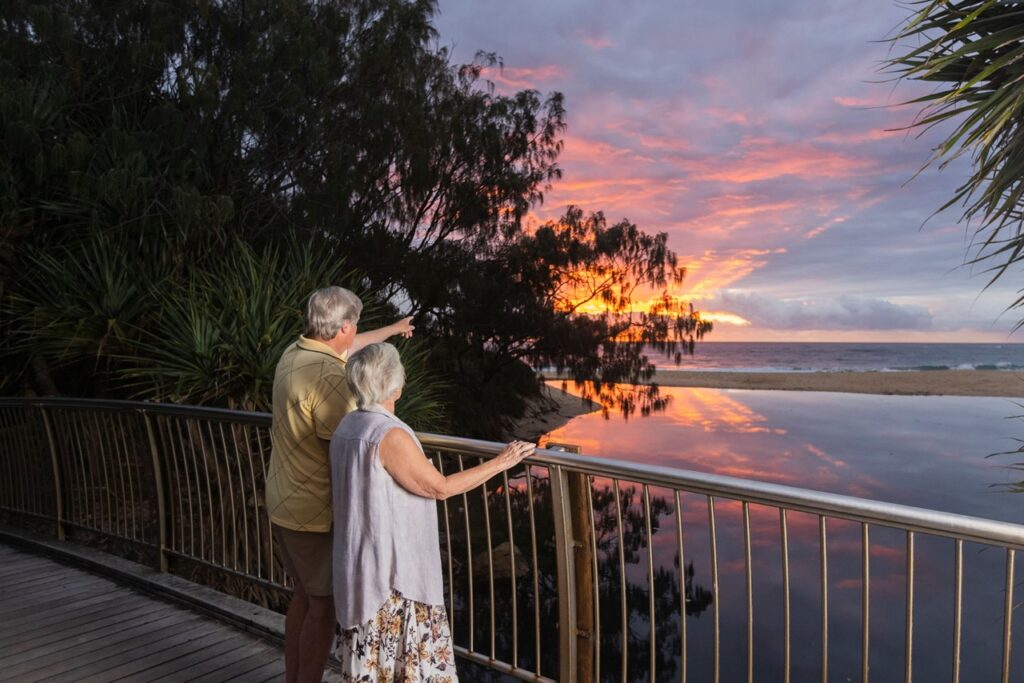 An elderly couple stands on a boardwalk, watching a vibrant sunset over a body of water, surrounded by trees.