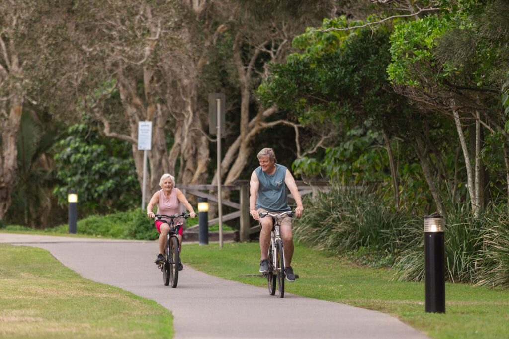 Two people are riding bicycles on a paved path in a park setting surrounded by trees and grass.