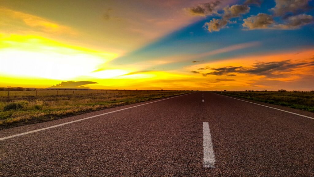 An empty road stretches towards the horizon under a vibrant sunset sky with scattered clouds.