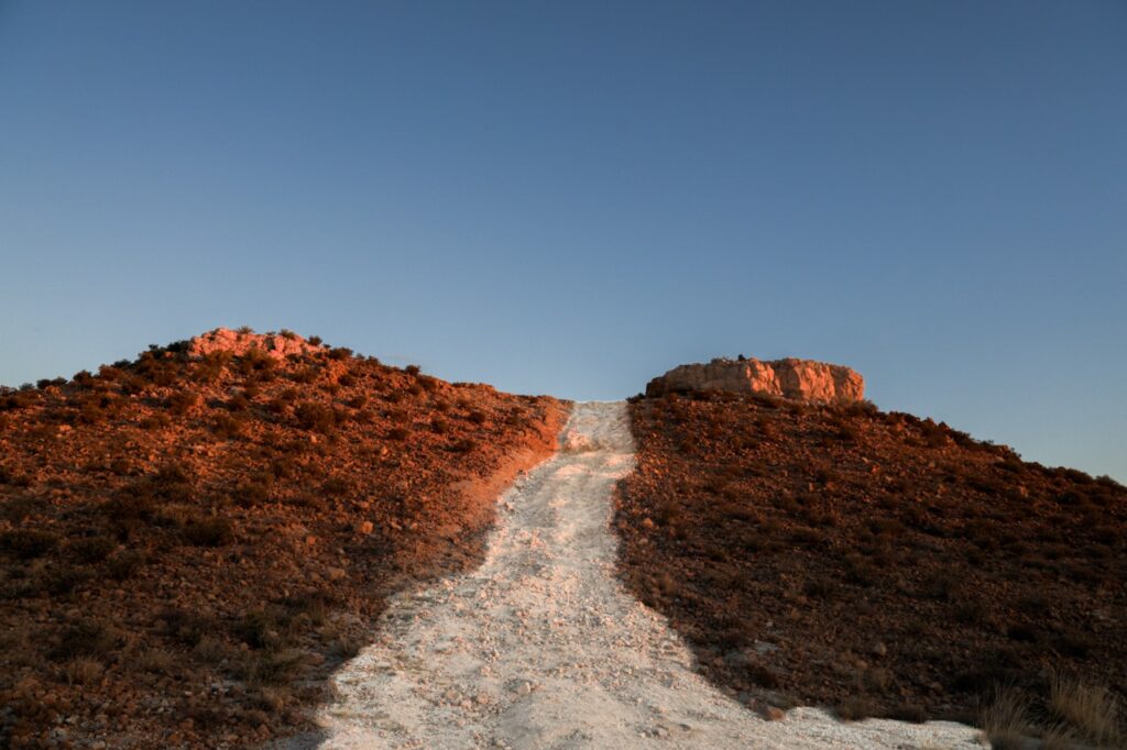 A dirt path leads up between two rocky hills under a clear blue sky during a sunset.