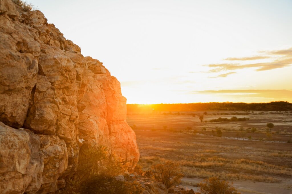 Sun setting over a rocky cliff with a dry landscape and sparse vegetation in the background.