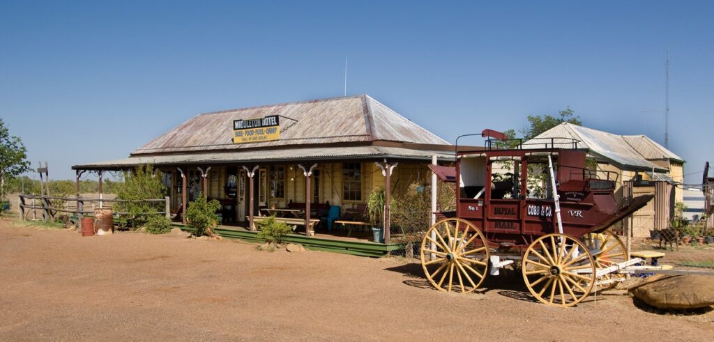 A rustic building with a tin roof and porch sits under a clear sky. A vintage stagecoach is parked on the dusty ground in front.