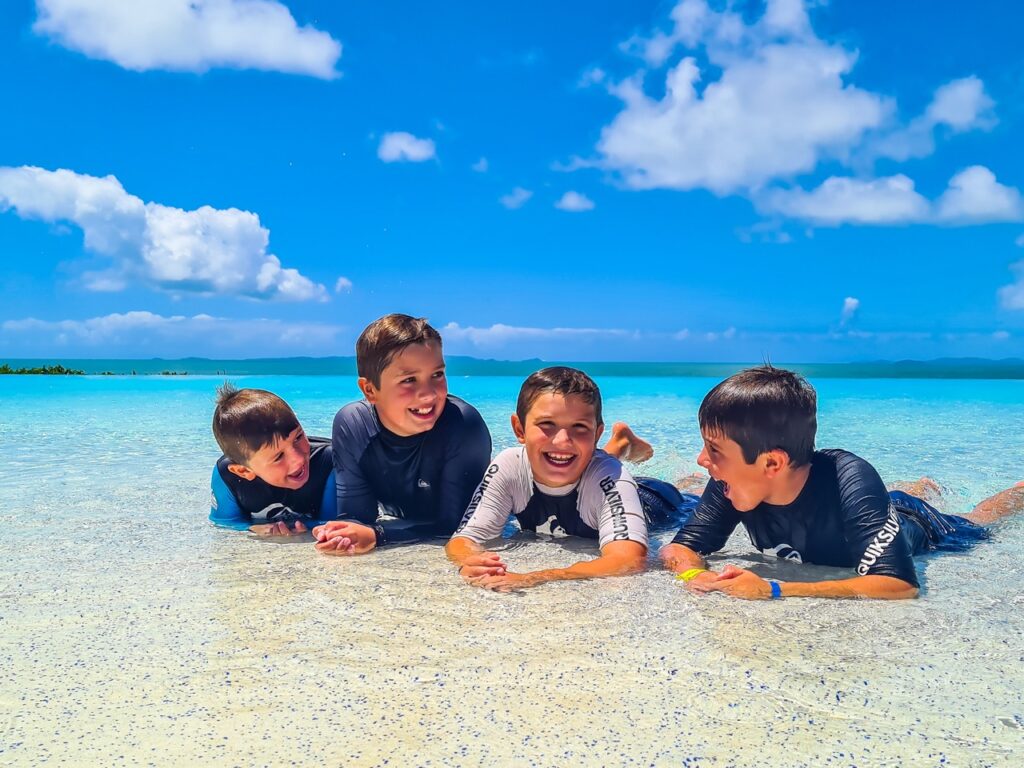 Four boys in swimsuits lying on shallow clear water, with blue sky and ocean in the background, smiling and talking.