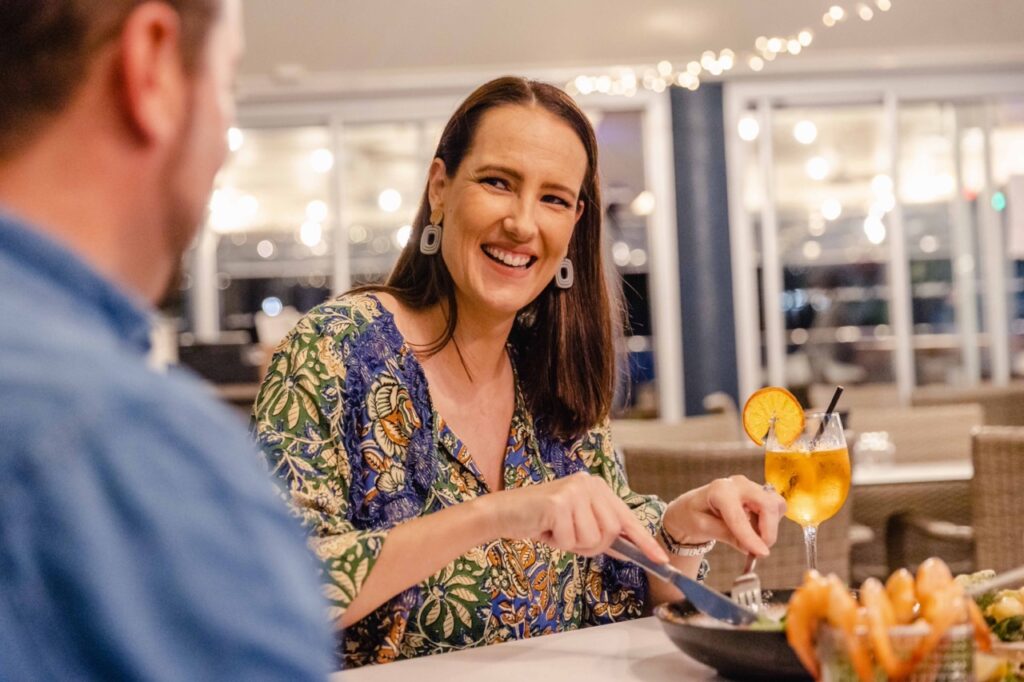 A woman smiles while dining with a man in a restaurant. She is cutting food on her plate, and a cocktail with an orange slice is on the table. Bright lights are visible in the background.