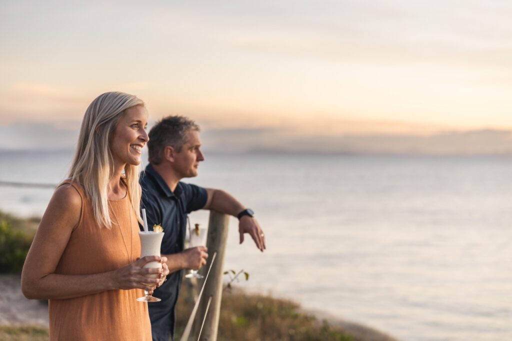 Two people standing by the water at sunset, one holding a drink, both looking towards the horizon.