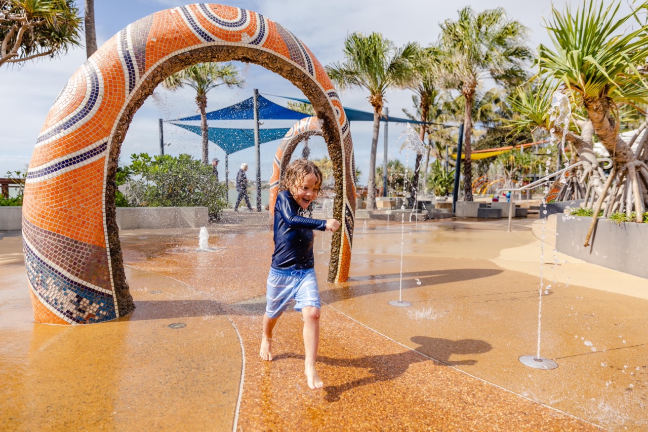 Child running through a water play area with colorful arches and palm trees in the background.