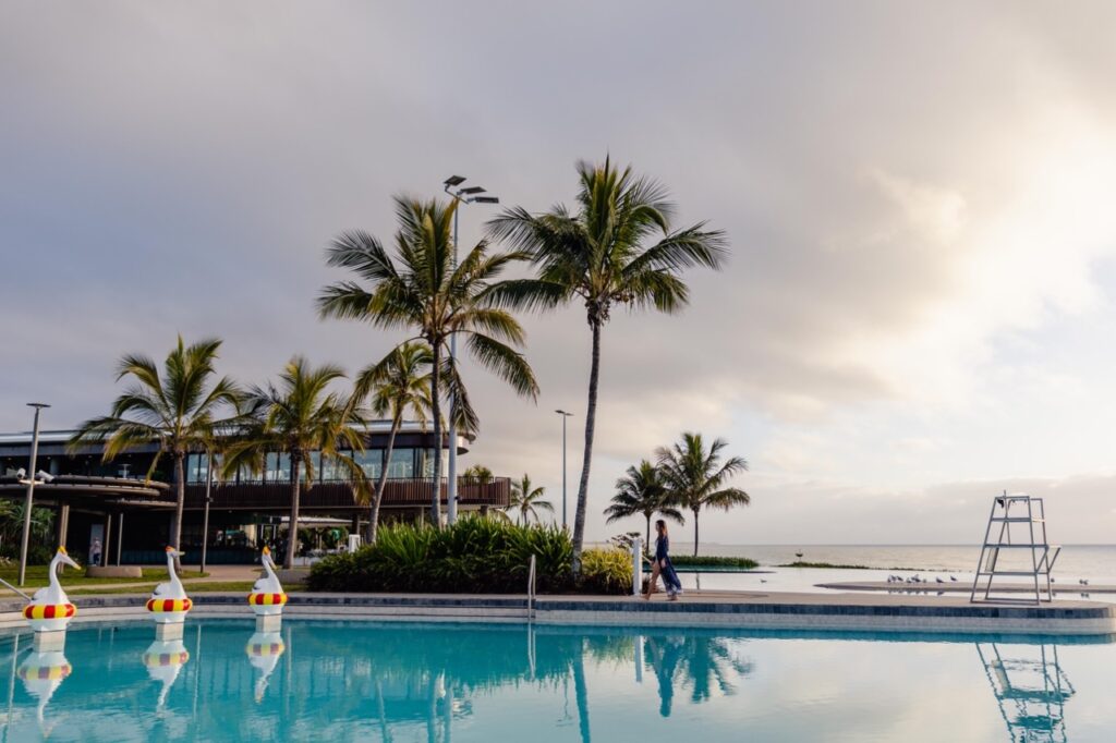 Outdoor pool scene with palm trees, a modern building, swan pool floats, and ocean view in the background. Two people walk on the pathway.