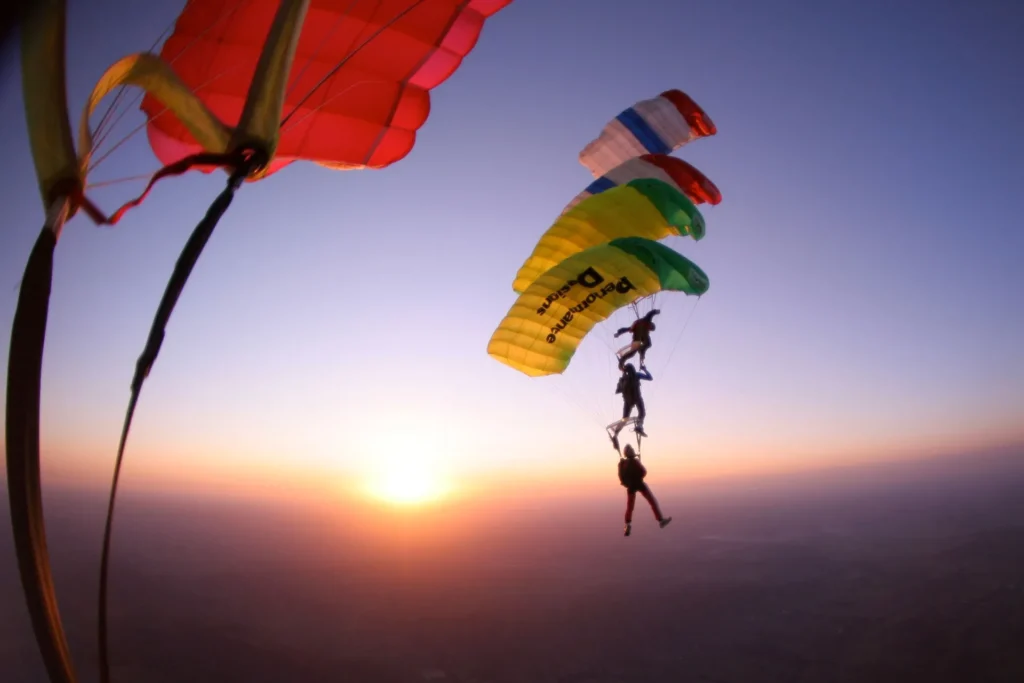 Three parachutists in colorful canopies perform a stacked formation during sunset, with the sky and horizon in the background.