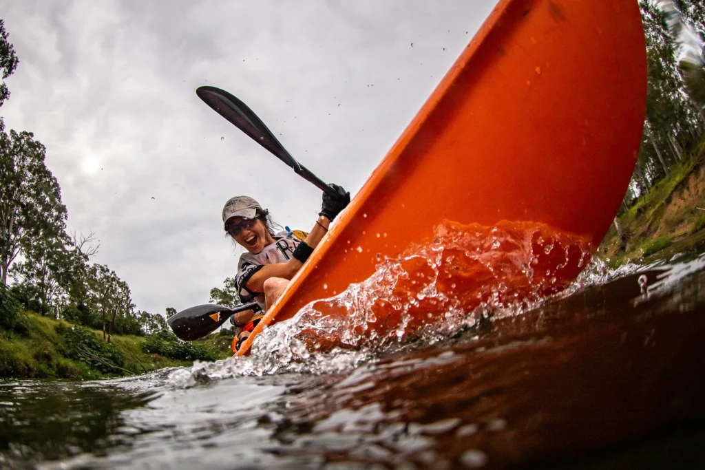 A person in a cap paddles a bright orange kayak through splashing water on a cloudy day, surrounded by trees.