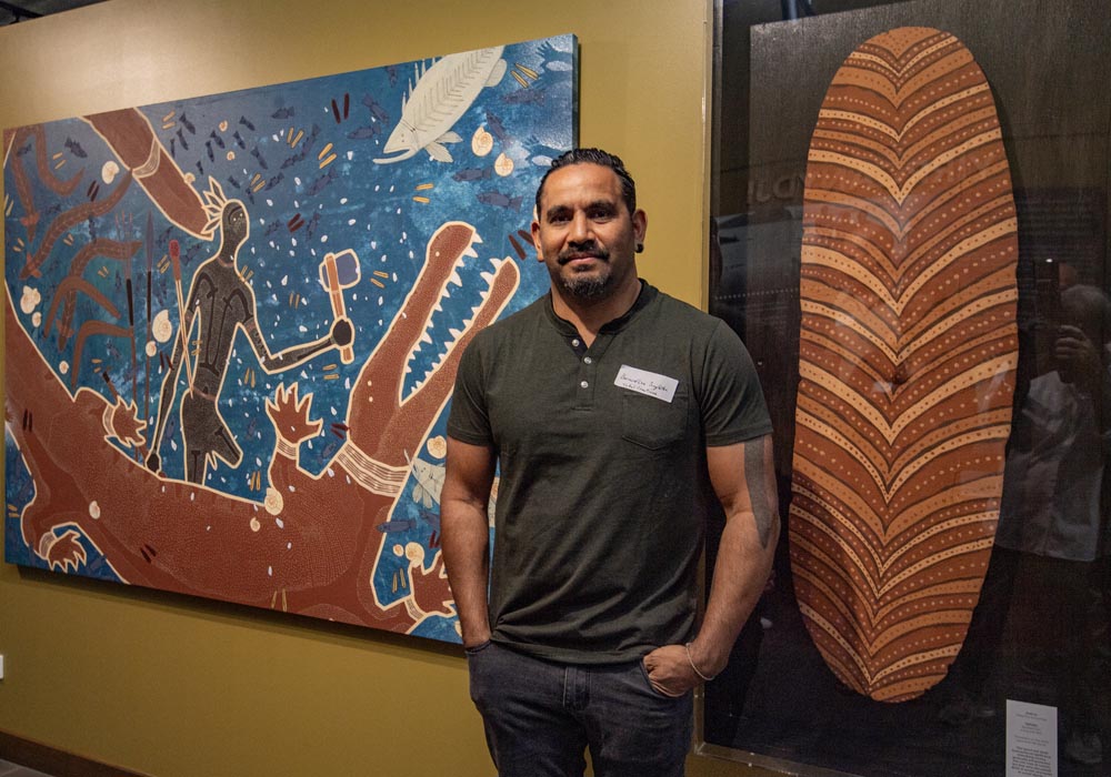 A man stands in front of two large, colorful Indigenous art pieces depicting a crocodile and traditional patterns.