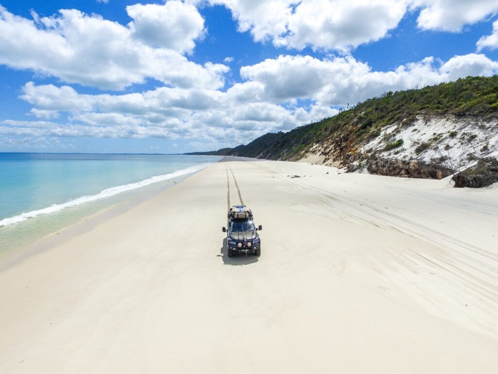 A black SUV drives along a vast, sandy beach with the ocean on the left and a hill with vegetation on the right under a partly cloudy sky.