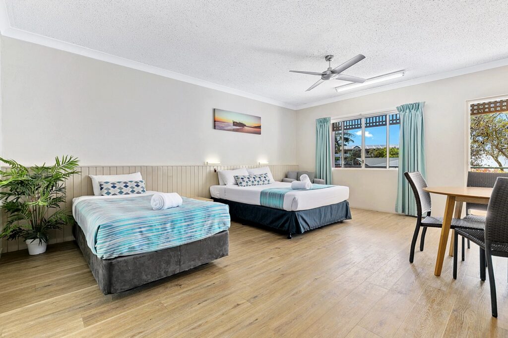 A spacious hotel room with two beds, wooden flooring, a ceiling fan, and a large window with pale blue curtains. A small table with chairs and a potted plant are also present.
