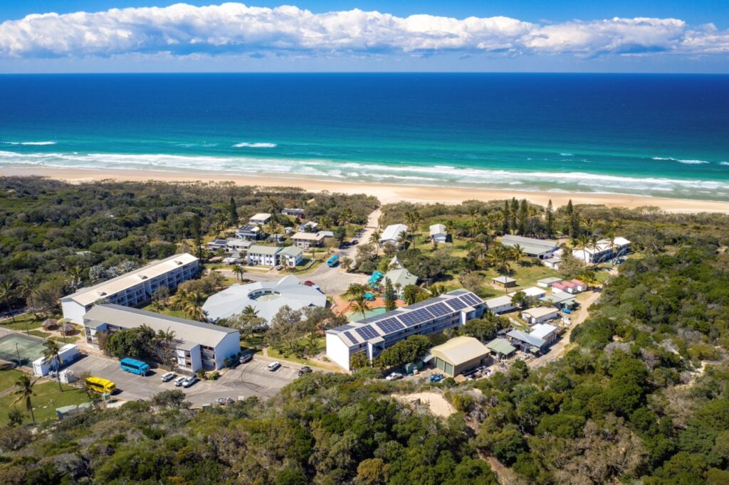 Aerial view of a coastal resort with multiple buildings surrounded by greenery. Solar panels are visible on some roofs. The ocean and beach are in the background under a partly cloudy sky.