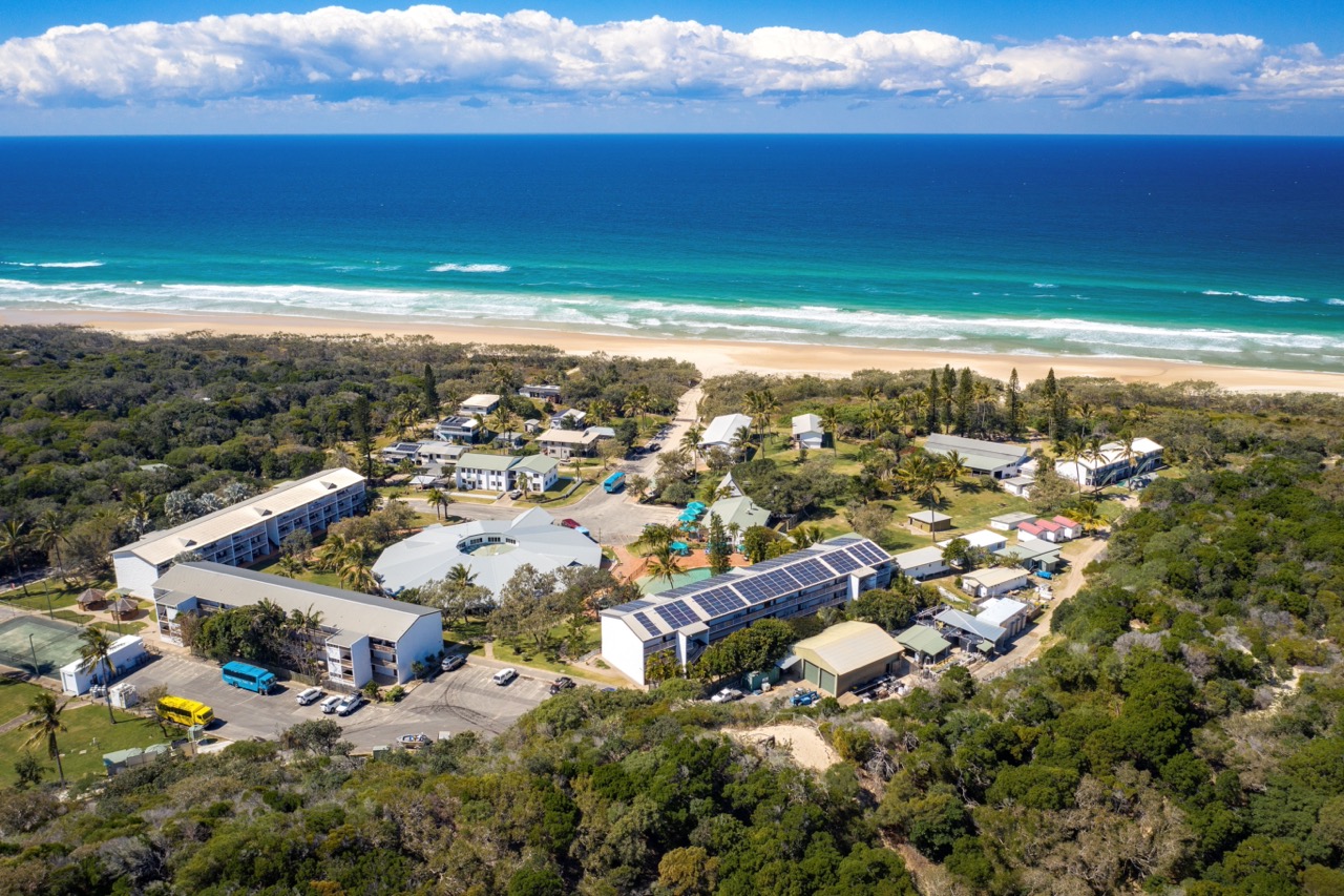 Aerial view of a coastal resort with multiple buildings surrounded by greenery. Solar panels are visible on some roofs. The ocean and beach are in the background under a partly cloudy sky.