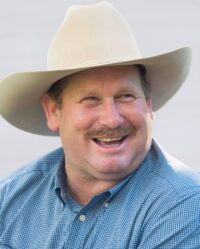 A man wearing a cowboy hat and a blue checkered shirt smiles while looking into the distance.