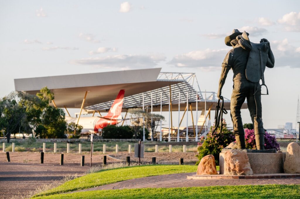 Statue of a man with a hat and whip in the foreground, with a large modern building and an airplane tail in the background.